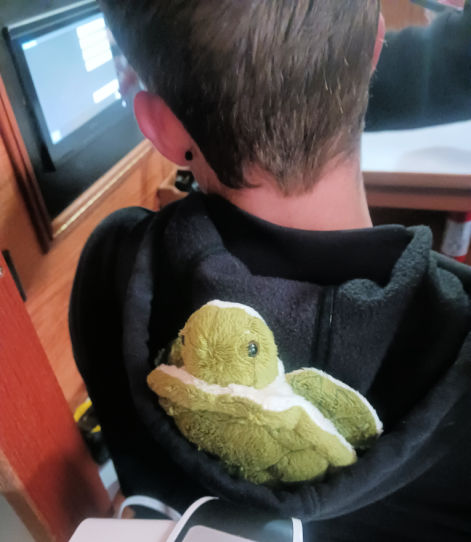 stuffed animal turtle in young man's hoodie hood while sitting at nav desk in sailboat 