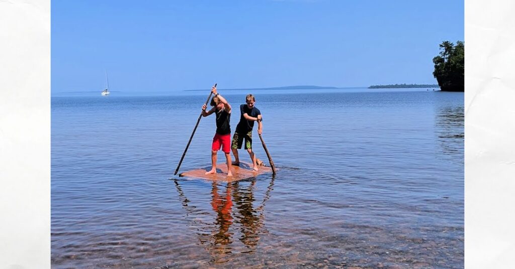 teen boys on a raft moving it along using long sticks, sailboat anchored in the background, calm lake superior 