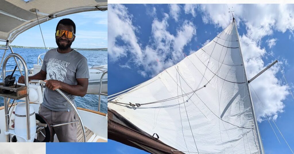 young man at wheel of sailboat, mainsail up with partly cloudy sky in background