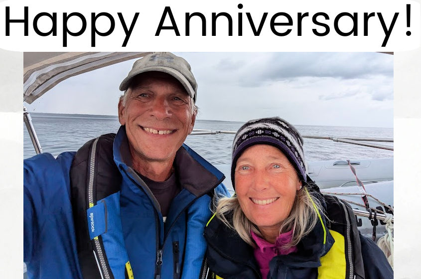 smiling couple on sailboat with water in background, life vests on, hats, jackets 