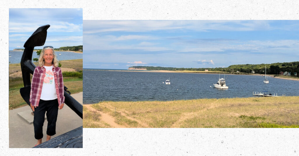 view of Grand Marais harbor from the town with sailboat and fishing boats at anchor or on mooring, woman by large anchor overlooking the bay 