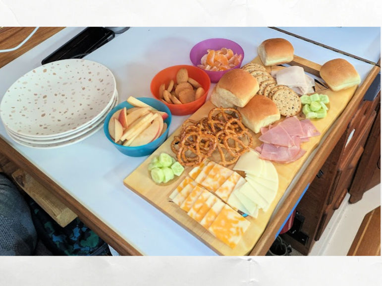 charcuterie board spread for sandwiches, apples, cukes, pretzels in sailboat cabin with plates ready to serve 