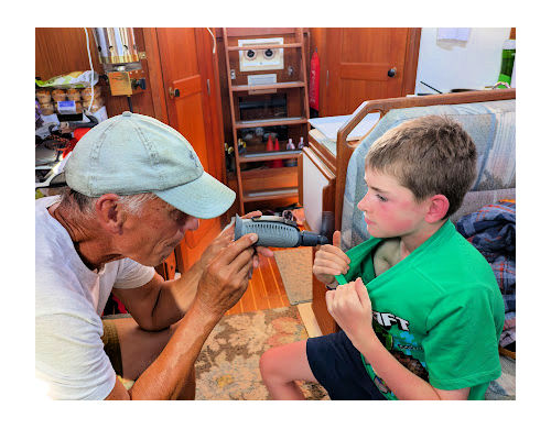grandpa using hand held fan to cool grandson's sunburn on his chest, they are in sailboat cabin 
