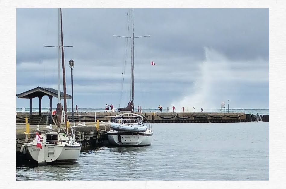 sailboats tied up to wall, big wave crashing over breakwater in distance, people on breakwater getting wet from the wave
